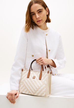 Žena v bílé bundě držící béžovou tkanou kabelku s hnědými koženými rukojeťmi, sedící na pozadí jednobarevné bílé stěny.