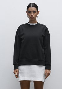Sudadera de algodón negro con cuello redondo, hombros caídos y puños acanalados. Llevada sobre un vestido mini blanco, que presenta un corte holgado y una textura suave.