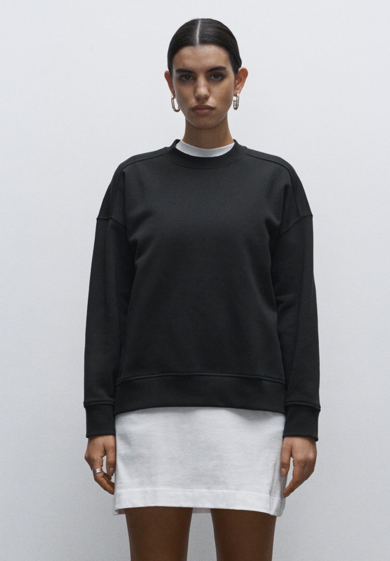 Sudadera de algodón negro con cuello redondo, hombros caídos y puños acanalados. Llevada sobre un vestido mini blanco, que presenta un corte holgado y una textura suave.