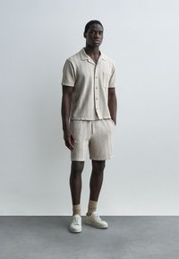 Homme debout contre un mur gris, portant une chemise à manches courtes boutonnée beige texturée, un short assorti, des chaussettes beiges et des baskets blanches.