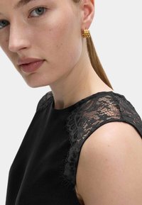 Schwarzes Kleid mit Spitze an den Schultern; glatte Textur mit taillierter Passform; Nahaufnahme von Schulter und Ohr mit einem goldenen Ohrring.