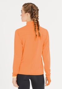 Oranssi urheilullinen pitkähiha, jossa on korkea kaulus, sileä pinta, peukalolenkit ja sauma selkäpuolella. Istuvuus on kapea.