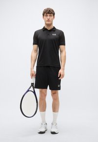 Juoda polo marškinėliai su logotipu, derinami su juodais šortais. Laiko teniso raketę, dėvi baltas kojines ir sportinius batelius. Paprastas dizainas.