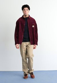 Bordeaux corduroy skjorte med knapper over en sort skjorte, parret med tan bukser og brune sko, stående på et lyseblåt gulv.