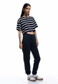 Marineblå cropped t-shirt med hvide vandrette striber; parret med mørke joggers. Modellen bærer hvide sneakers med sorte detaljer og lyse sokker.