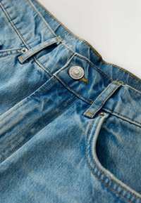Gros plan sur la ceinture d'un jean en denim bleu avec un bouton en métal, des passants de ceinture et des coutures de la poche avant sur un fond clair.