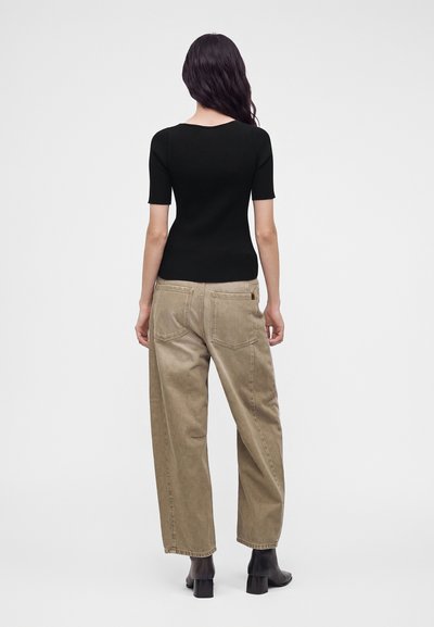 Femme aux longs cheveux foncés portant un haut côtelé noir à manches courtes, un pantalon beige à jambes larges et des bottines noires à talons, vue de dos.