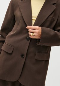 Brun blazer med notched lapels, to frontlommer, enkelt svart knappehold, og en lysere gul topp under. Gullring båret på hånden.