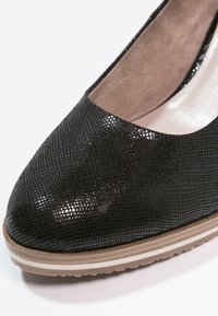 Svart texturerat läderpumpsko med en låg, rund tån. Har en ljusfärgad gummisula med ett subtilt vitt streck. Fodrad insida.