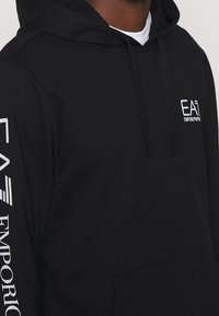 Sudadera negra hecha de una tela suave, con un bolsillo frontal, capucha con cordón y logotipos en blanco: "EA" en el pecho y "EMPORIO" en la manga.