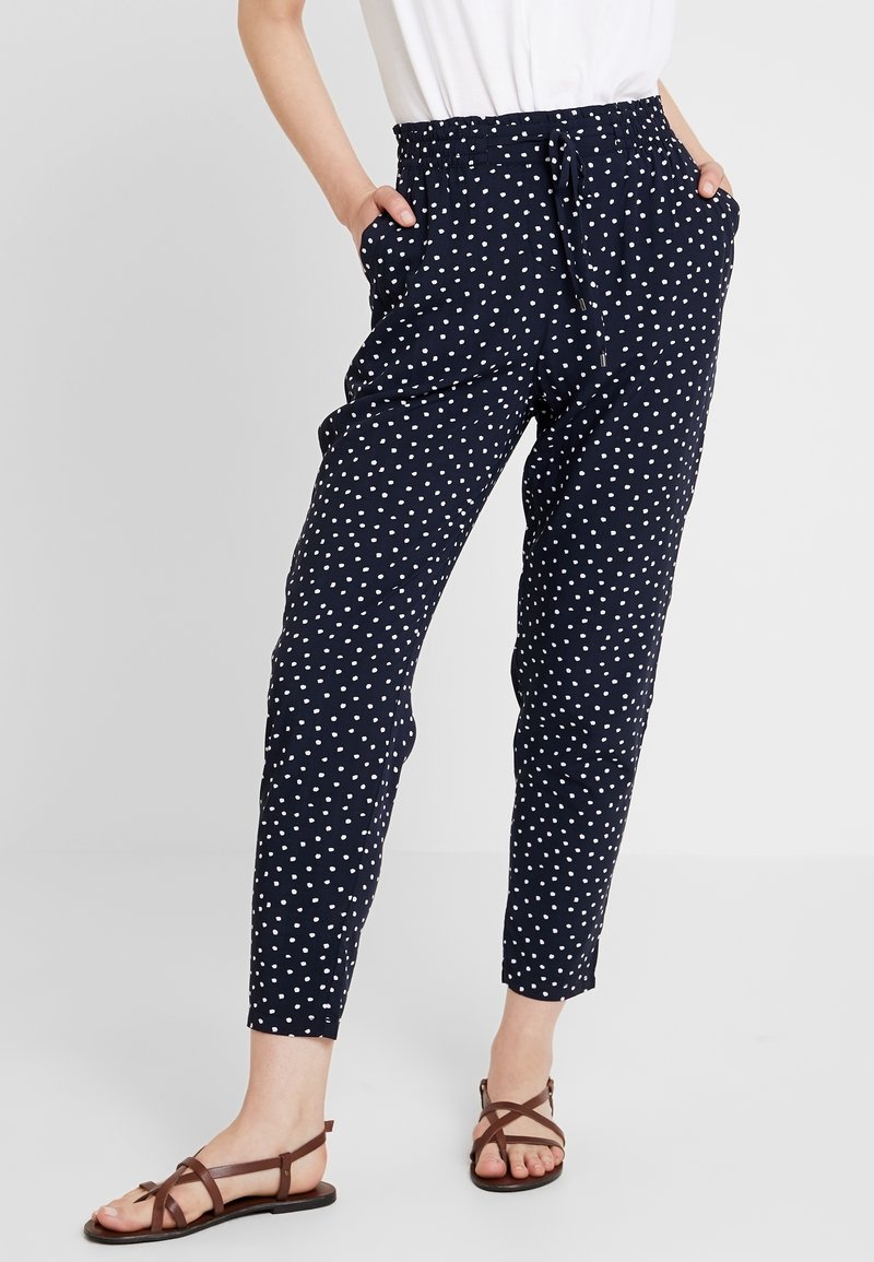 Femme portant un pantalon bleu marine fuselé à pois blancs, un haut blanc et des sandales plates marron à lanières, les mains dans les poches.