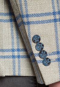 Beige jacket with a blue plaid pattern, textured fabric, and four round blue buttons on the cuff. Tailored design with detailed stitching.