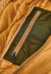 Geëlektrificeerde gele jacks aan de binnenkant met een groene ritszak en een beige rits. De stof heeft een subtiel ruitjespatroon en een gladde textuur.