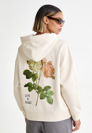 Vrouw draagt een crèmekleurige hoodie met bloemenprint en de tekst "LOVE & ROSES" op de rug, kort steil haar en donkere zonnebril, zijwaarts gezicht.