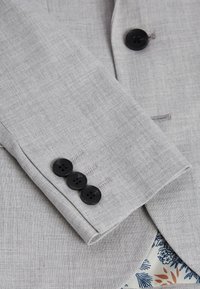 Manica di giacca in suit grigio chiaro con quattro bottoni neri, texture liscia e motivo intrecciato sottile. Fodera con un design floreale.