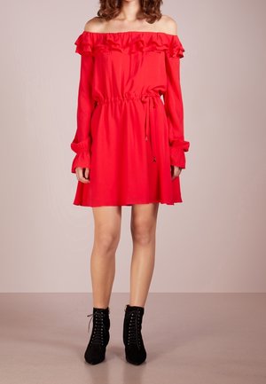 Femme portant une robe rouge épaules dénudées avec des volants et des manches longues, associée à des bottines noires à lacets, debout sur un sol uni.