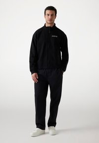 Black zip-up jacket with ribbed cuffs, featuring a logo. Paired with loose black pants and white sneakers on a neutral background.
