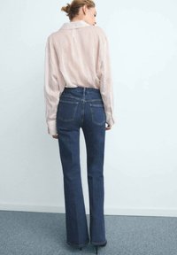Jeans in denim scuro a vita alta e svasati, con orlo grezzo e tasche posteriori, abbinati a una camicia a maniche lunghe a righe rosa chiaro.