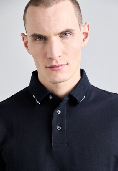 Polo shirt bleu marine foncé en tissu texturé, avec un col classique, trois boutons et un logo blanc sur le col.