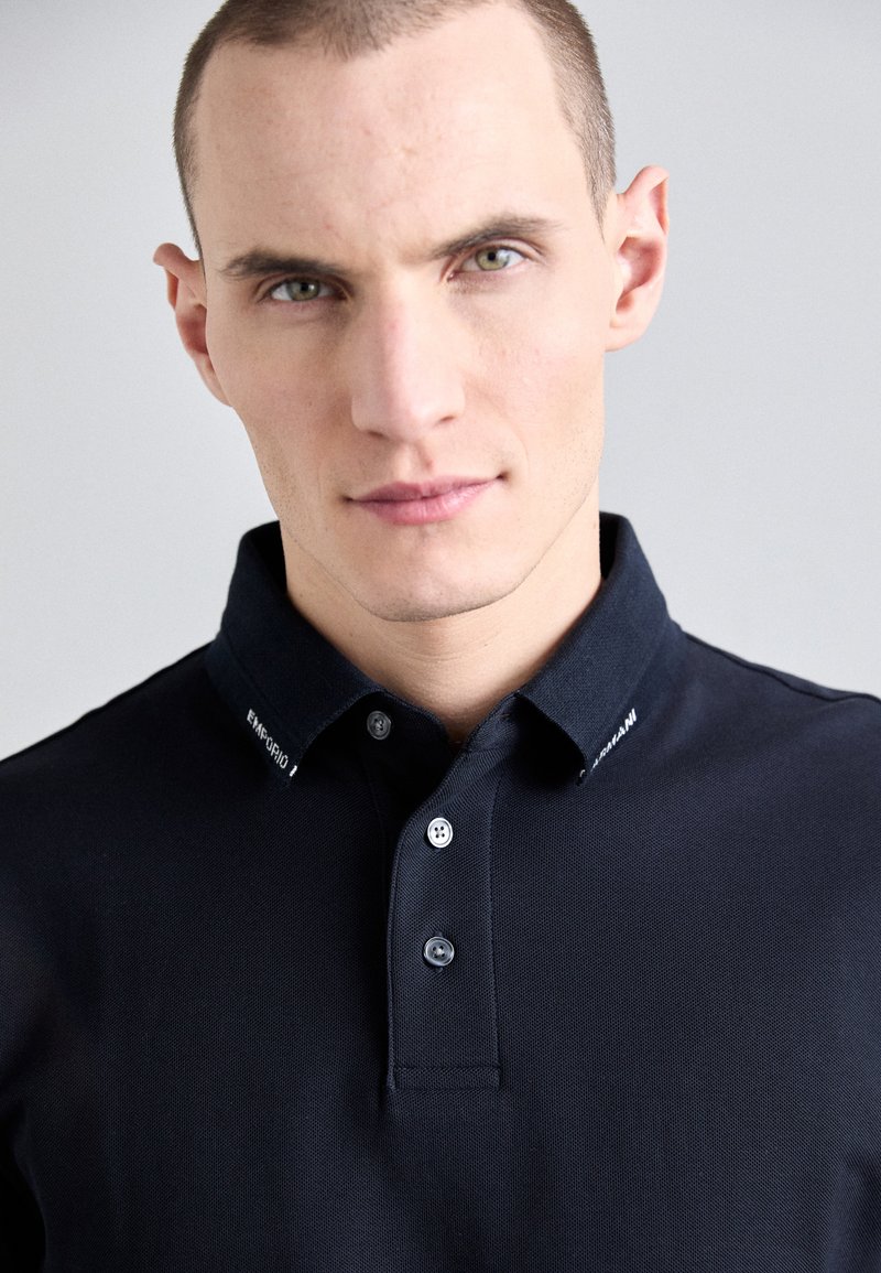 Mörk marinblå polo skjorta med en texturerad tyg, med en klassisk krage, tre knappar och vit branding på kragen.