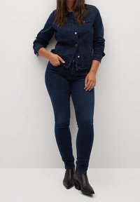 Mujer con chaqueta de mezclilla azul oscuro y jeans, combinados con botines negros, posando frente a un fondo liso de color claro.