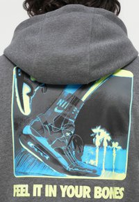 Close-up de um hoodie cinza escuro com um gráfico azul e amarelo de ténis de corrida e palmeiras, com o texto "SENTE-TE ISSO NOS OSSOS."