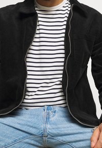 Svart kordflöjlsjacka med dragkedja, bärs över en vit och marinrandig t-shirt. Blå denimjeans syns, vilket visar en avslappnad passform.