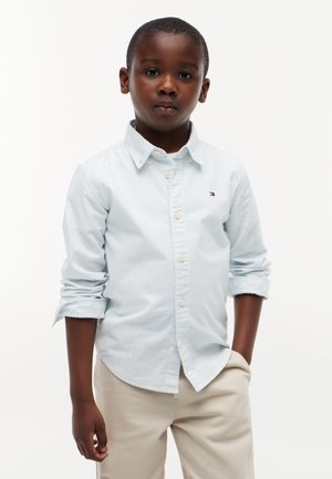 Garçon aux cheveux courts portant une chemise bleu clair à boutons avec les manches retroussées et un pantalon beige, debout avec une main dans la poche.
