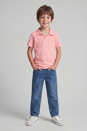 Ragazzo giovane sorridente in piedi con le mani nelle tasche, indossa una polo rosa, jeans blu e sneakers bianche su sfondo semplice.