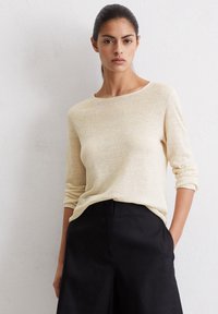 Jasno beżowy sweter z długim rękawem wykonany z miękkiego, teksturowanego materiału wełnianego. Posiada okrągły dekolt i luźny krój, w zestawie z czarnymi spodniami.