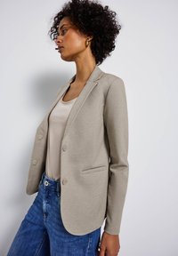 Blazer entallado beige con solapas muesca, dos botones y un bolsillo en el pecho; combinado con una blusa clara y jeans azules.