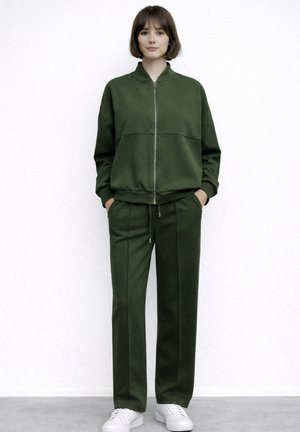 Giovane donna che indossa una giacca con zip verde scuro e pantaloni abbinati, in piedi con le mani nelle tasche, scarpe da ginnastica bianche, sfondo neutro.