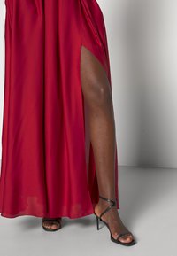 Abito rosso fluente con un'alta spaccatura. Tessuto morbido e leggero che si adagia fino a terra. Sandali neri con tacco e fasce sottili che completano l'outfit.