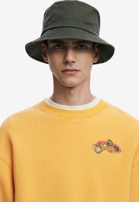 Gul sweatshirt med rund halsudskæring og et lille broderet design på brystet, parret med en mørkegrøn bøttehat.