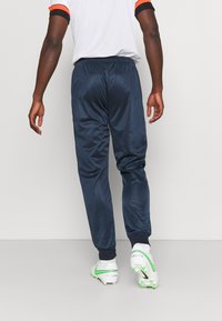 Pantaloni sportivi blu navy con texture liscia, vita elasticizzata e gambe affusolate, abbinati a scarpette da calcio bianche con accenti verdi.