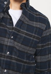Camicia flanella a quadri navy e grigia con colletto button-down. Presenta una tasca sul petto e bottoni, evidenziando un tessuto testurizzato.