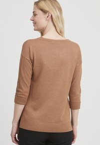 Brązowy sweter dzianinowy z rękawami o długości trzech czwartych i podwiniętymi mankietami, z guzikami wzdłuż szwów rękawów. Miękka tekstura i luźny krój.