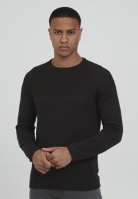 Chemise noire à manches longues avec une texture côtelée et un panneau lisse contrastant sur la poitrine. Encolure ronde, design ajusté.