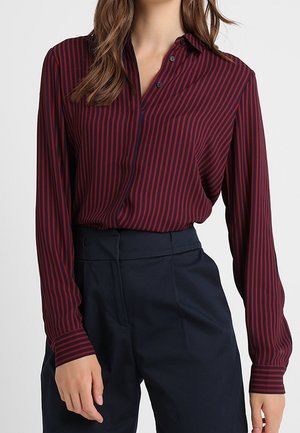 Mujer con una blusa de manga larga a rayas verticales rojas y negras metida en pantalones negros de talle alto, frente a un fondo sencillo.