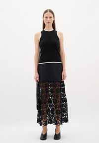 Black sleeveless top with white trim, layered over a long black lace skirt featuring floral patterns, paired with black heeled shoes.
