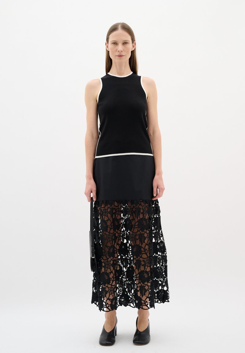 Black sleeveless top with white trim, layered over a long black lace skirt featuring floral patterns, paired with black heeled shoes.