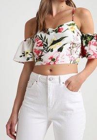 En kvinna som bär vita högmidjade jeans och en vit blommig crop top med avskurna puffärmar och tunna axelband.