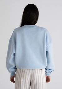 Sudadera oversized de color azul claro hecha de tela suave, con hombros caídos y puños acanalados, combinada con pantalones rayados en beige y blanco.