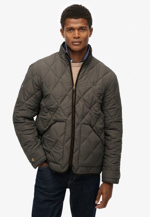 Hombre vestido con una chaqueta acolchada de oliva oscuro sobre un suéter beige y una camisa de mezclilla azul, de pie con una mano en el bolsillo frente a un fondo blanco.