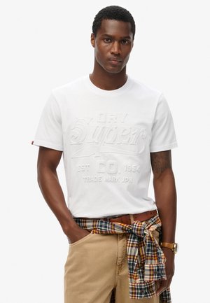 Camiseta de algodón blanca con texto en relieve, combinada con pantalones caqui y una camisa a cuadros multicolor atada a la cintura.