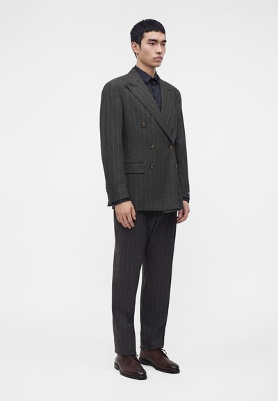 Homme debout portant un blazer croisé à fines rayures gris foncé, un pantalon assorti, une chemise noire et des chaussures habillées en cuir marron.