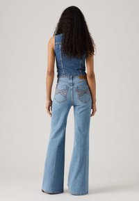 Pikalt pikkade lokkis juustega naine, seljas ilma varrukateta denim vest ja kõrge vöökohaga helesinised laiad teksad, seismas ühtlase tausta ees.