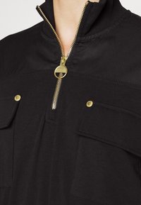 Black pullover with a quarter-zip closure and gold zipper. Features two front pockets with snap buttons and a smooth, textured fabric.