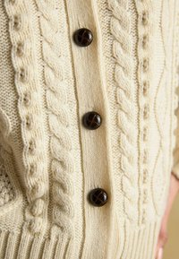 Krémový pletený kardigán s károvanými vzormi a štyrmi tmavohnedými gombíkmi s krížovým vzorom na prednej strane. Textúrovaná tkanina.