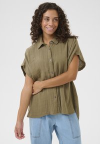 Olive green, short-sleeved button-up shirt with a textured fabric, featuring a collar and slightly dropped shoulders, paired with light blue trousers.